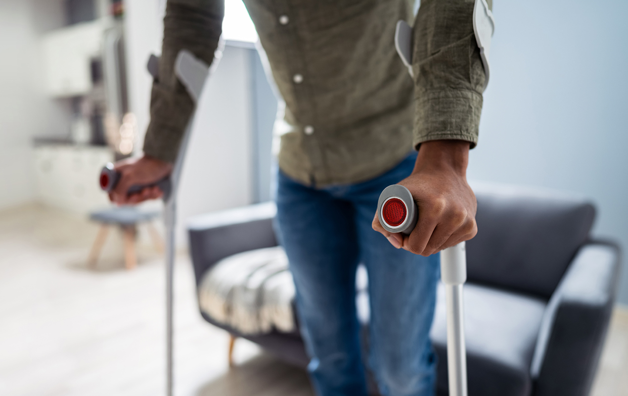 Person steht in einem Wohnzimmer und stützt sich auf Krücken. Im Hintergrund sieht man unscharf eine Couch.